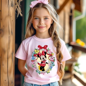 Peut inclure: Une jeune fille porte un t-shirt rose clair avec un motif de Minnie Mouse. Le motif repr&eacute;sente Minnie Mouse dans une robe &agrave; pois rouges et un n&oelig;ud, entour&eacute;e de fleurs. Elle porte &eacute;galement un bandeau rose avec un n&oelig;ud.
