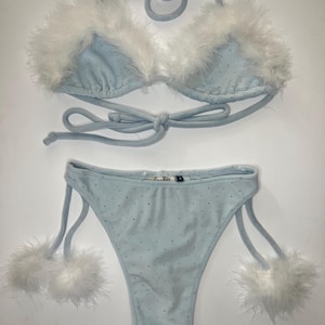 May include: A light blue bikini with white feather trim. The top is a triangle style with adjustable straps and the bottom is a high-cut style with ties on the sides. The bikini is made of a soft, stretchy material and has a sparkly finish.