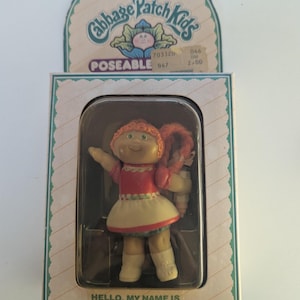 Könnte beinhalten: Cabbage Patch Kids Phoebe Kim Puppe in Originalverpackung. Die Puppe hat rotes Haar, ein rotes und weißes Kleid und blaue Schuhe. Die Verpackung enthält das Cabbage Patch Kids Logo und den Text "POSEABLE". Der Text "HELLO, MY NAME IS PHOEBE KIM" ist ebenfalls sichtbar.