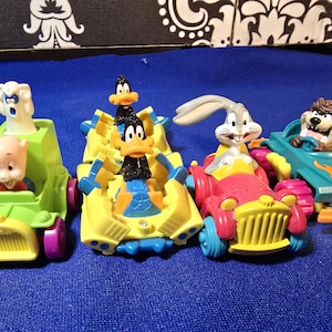 May include: Four colorful toy cars featuring Looney Tunes characters. The cars are in shades of green, yellow, red, and teal, with characters like Porky Pig, Daffy Duck, Bugs Bunny, and the Tasmanian Devil in the driver's seats.