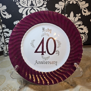 Può includere: Un piatto decorativo con centro bianco e bordo bordeaux con dettagli dorati. Il piatto presenta le parole "Ruby 40th Anniversary" in lettere dorate ed è esposto su un supporto trasparente.