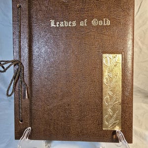 以下が含まれることがあります： 「Leaves of Gold」のタイトルが金色の文字で書かれた茶色のフォトアルバム。アルバムは、テクスチャード加工された表紙と、葉のデザインが施された金色の装飾アクセントが特徴です。茶色のコードでアルバムを閉じます。