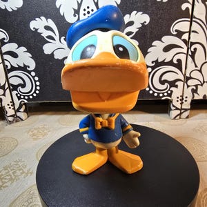 May include: A Donald Duck figurine. The figure has a large orange beak, blue eyes, and a blue sailor hat. He is wearing a blue sailor shirt with a yellow bow tie and orange feet. The figurine is standing on a black circular base.