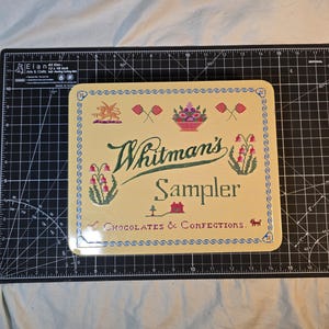 Könnte beinhalten: Eine quadratische, cremefarbene Whitman's Sampler Schokoladen- und Konfektdose mit Kreuzstich-Stickmustern eines Drachen, Blumen, eines Korbs und eines Hauses sowie grünem Text auf einer schwarzen 30,48 x 45,72 cm großen Schneidematte.