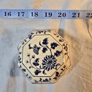 Könnte beinhalten: Achteckige Keramik-Schmuckschatulle mit cremefarbenem Grund und blauem Blumendesign. Der Deckel zeigt eine zentrale Blume mit umgebenden Blättern und kleinere Blumenmuster an den Rändern. Die Box ist etwa 18 cm lang.