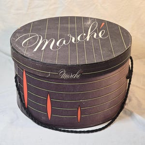 May include: A vintage round hat box with the word "Marché" in white script on the lid. The box is primarily dark brown with vertical gold stripes and orange accents. It has a black rope handle.