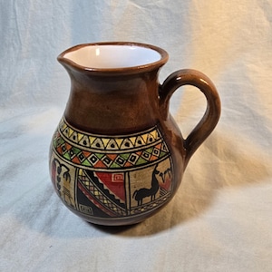 May include: A brown ceramic pitcher with a white interior and a single handle. The exterior features a hand-painted band with geometric patterns in green, yellow, and red, along with a black silhouette of a llama and other abstract shapes.