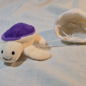 May include: A plush sea turtle toy with a cream-colored body and a vibrant purple shell. The turtle is attached by a thin white cord to a separate, hollow, white plush dome-shaped piece that resembles a small pouch or a matching accessory.
