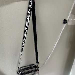 Op de afbeelding: Een doorzichtige crossbodytas met een zwarte rand en een zwarte band. De band heeft witte tekst met de tekst "KOZUZU". De tas heeft een rechthoekige vorm met een ritssluiting.