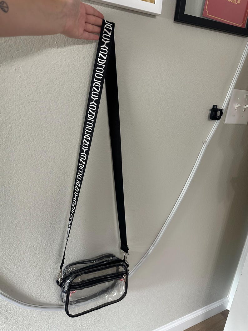 Op de afbeelding: Een doorzichtige crossbodytas met een zwarte rand en een zwarte band. De band heeft witte tekst met de tekst "KOZUZU". De tas heeft een rechthoekige vorm met een ritssluiting.