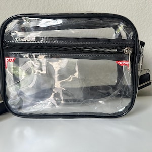Op de afbeelding: Een transparante, rechthoekige crossbodytas met zwarte randen en een ritssluiting. De tas heeft een zwarte verstelbare riem en een klein rood-wit logo. Hij ligt op een wit oppervlak.