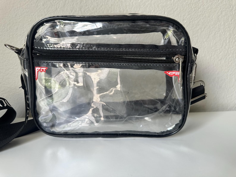 Op de afbeelding: Een transparante, rechthoekige crossbodytas met zwarte randen en een ritssluiting. De tas heeft een zwarte verstelbare riem en een klein rood-wit logo. Hij ligt op een wit oppervlak.