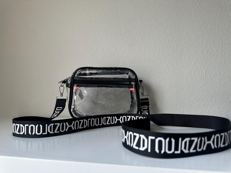 Op de afbeelding: Transparante crossbodytas met zwarte rand en een zwart-witte band. De tas heeft een ritssluiting aan de bovenkant en een rode accent. De band heeft witte tekst. De tas staat op een wit oppervlak.