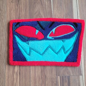 May include: A rectangular rug featuring a cartoon face design. The rug has a red border, with a dark blue background. The face has red eyes, a light blue mouth, and black accents. The rug is placed on a wooden floor.