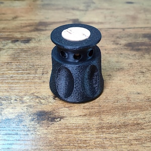 Può includere: Un oggetto nero cilindrico con una superficie strutturata e una parte superiore piatta. L'oggetto ha una serie di piccoli fori attorno al centro e un design circolare sui lati. C'è un adesivo bianco sulla parte superiore.