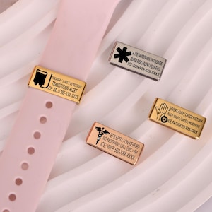 Könnte beinhalten: Ein rosafarbenes Smartwatch-Armband mit mehreren medizinischen Metallanhängern. Die Anhänger sind gold-, silber- und kupferfarben und mit medizinischen Informationen und Symbolen graviert. Die Anhänger enthalten Warnhinweise für Transfusionen, Allergien und Epilepsie.