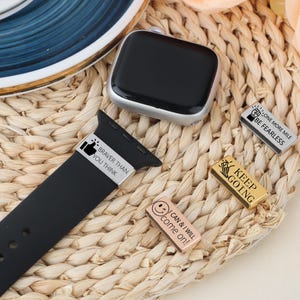 May include: A black smartwatch with a silver case and a black band, alongside several decorative watch band charms. The charms are silver and gold, with motivational phrases like "Braver Than You Think" and "Keep Going."