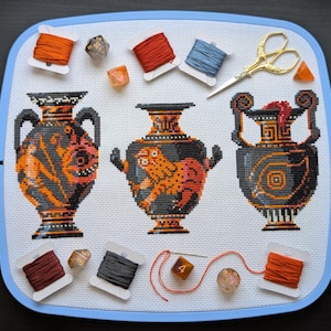 May include: Cross-stitch design showing three black and orange Greek vases on white fabric, with orange, red, and blue thread spools, dice, and gold scissors. The project is in a blue embroidery hoop.