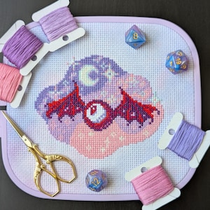 May include: Cross-stitch project featuring a fantasy creature with a large eye, wings, and a crescent moon, in shades of purple, pink, and red. Gold scissors, spools of pink and purple thread, and blue dice surround the project on a light purple frame.