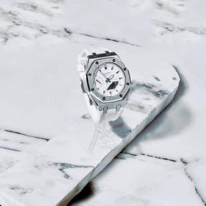 Puede incluir: Un reloj de pulsera blanco y plateado con una correa blanca. El reloj tiene un bisel plateado y una esfera blanca con manecillas negras y una pantalla digital. Se muestra sobre una superficie de mármol blanco.