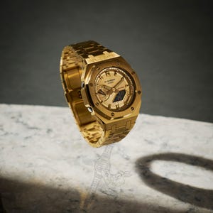 May include: A gold-toned G-Shock Casio watch with a matching gold band. The watch face displays multiple dials and a digital display. The watch is set against a neutral background, showcasing its metallic finish and intricate design.