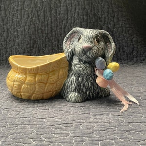 May include: A gray ceramic bunny holding a string of pastel colored Easter eggs. The bunny is sitting next to a yellow woven basket.