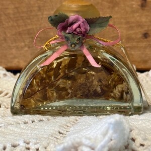 May include: A clear, square-shaped glass bottle filled with dried flowers. The bottle is topped with a wooden cap and adorned with a pink ribbon, a small pink flower, and green leaves. The bottle sits on a white, crocheted surface.