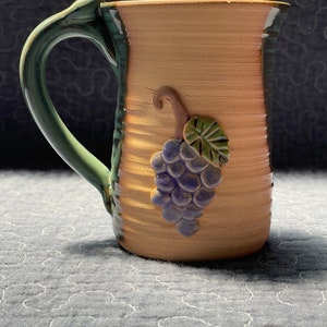 May include: A ceramic mug with a green handle and a brown body. The mug has a raised design of a grape vine with purple grapes.