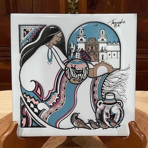 May include: A white ceramic tile with a colorful illustration of a woman in a blue and pink patterned dress holding a pot with a bird inside. The background features a white building with a blue sky and a bird flying overhead. The tile is signed "Tussedre '91".