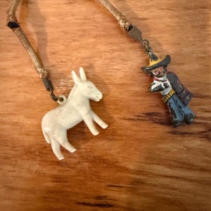 May include: A vintage keychain featuring two small charms: a white donkey and a miniature figure of a man in a sombrero and poncho. The charms are connected by a woven cord, set against a wooden background.
