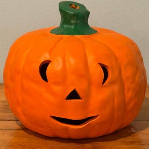 May include: A ceramic orange pumpkin with a green stem. The pumpkin has a carved face with crescent-shaped eyes, a triangular nose, and a curved mouth. This Halloween decoration is a classic festive item.