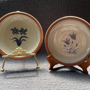 May include: Two ceramic plates with blue and white designs. The plate on the left has a floral design, while the plate on the right has a bird design. Both plates are on decorative stands.