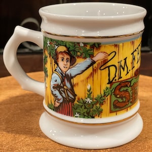 May include: White ceramic mug with a decorative handle and base. The mug features a colorful illustration of a young person in a hat, leaning against a wooden fence. The mug has gold trim and the text "D.M. FESS" is visible.