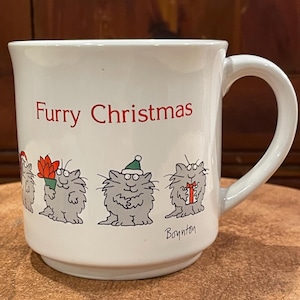 May include: White ceramic mug with a red "Furry Christmas" inscription and a line of four cartoon cats in grey, white, and red. The cats are wearing Santa hats and holding gifts. The mug is signed "Boynton" at the bottom.