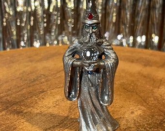 May include: A pewter figurine of a wizard holding a crystal ball. The wizard is wearing a pointed hat with a red jewel on top and a long robe. The figurine is detailed and has a realistic appearance.