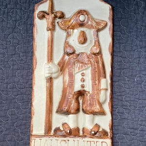 May include: A ceramic wall plaque featuring a cartoon-like figure holding a staff. The figure is wearing a brown coat, white shirt, and a hat. The plaque is white with brown accents and the word "HAUSHUTER" is written at the bottom.