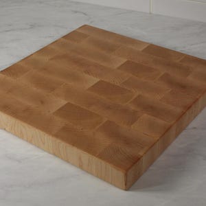 Peut inclure: Un bloc de boucher carré à grain de bout. La planche est en bois clair avec un motif rectangulaire. La planche est posée sur un plan de travail en marbre blanc et gris.