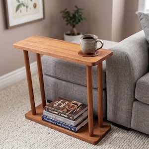 May include: A narrow rectangular wooden side table with a smooth finish, featuring four cylindrical legs and a lower shelf. The table holds a ceramic mug on a coaster and a stack of three books titled The Craft of Wood, Minimalist Homes, and A Short History of Nearly Everything.