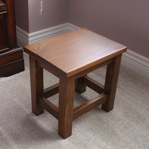 Puede incluir: Un taburete de madera marrón oscuro con un diseño cuadrado simple. El taburete tiene un acabado liso y pulido y una construcción robusta.