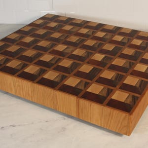 May include: A large, rectangular end-grain chopping board. The surface features a mosaic of light and dark wood squares, forming a geometric pattern. The sides are made of a lighter wood. It has a built-in handle on one side.