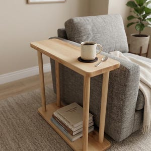 May include: A light-coloured wooden side table with a rectangular top and a lower shelf. The table has four cylindrical legs. A mug of coffee sits on the table, along with a spoon and a coaster. Several books are stacked on the lower shelf. The table is next to a grey sofa.
