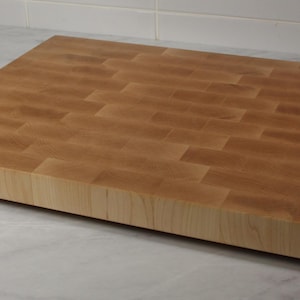 May include: A large, square, wooden chopping board made of maple wood. The board has a butcher block style construction with a smooth, light brown finish.