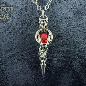 May include: A silver-toned pendant necklace with a red gemstone center. The pendant has a spiked bottom and is suspended from a chain. The text "Steampunk Garage" is in the upper left corner. The background is a dark, textured fabric.