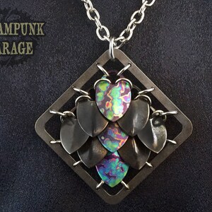 May include: A silver chain necklace with a square pendant featuring a cluster of iridescent, multi-colored, heart-shaped metal scales. The pendant is framed by a silver metal square.