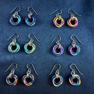 May include: A collection of colorful, handmade earrings. Each pair features a unique design of interlocking rings in shades of blue, purple, green, yellow, and pink. The earrings have silver-colored ear wires and are displayed on a dark blue fabric.