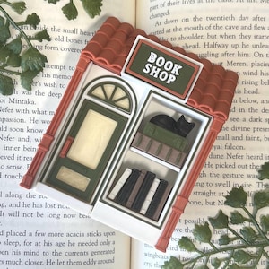 May include: A miniature book shop figurine with a terracotta-colored tiled roof, a green door, and a sign that reads BOOK SHOP. The shop front features a display window with shelves containing books and a small black cat figurine inside.