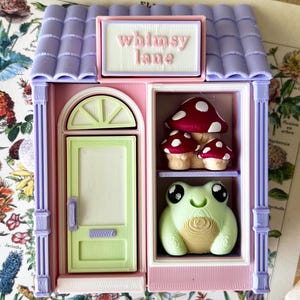 May include: A small, three-dimensional shop structure with a lavender roof and pink walls. A sign above the door reads "whimsy lane" in pink text. The shop features a light green door and a display window containing three red mushrooms and a light green frog.