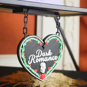 May include: A black heart-shaped sign with the words "Dark Romance" in white script, decorated with red roses, green leaves, and a small skull. The sign hangs from a black chain attached to a black metal shelf.