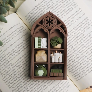 May include: A brown, arched, gothic-style miniature bookshelf with three levels. The shelves contain small decorative items including books, a potted plant, a jar, three white candles, a green frog figurine, and a black cat figurine.
