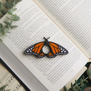 May include: An open book with a monarch butterfly bookmark. The bookmark is orange, black, and white. The book is surrounded by greenery and a small plant, with a musical score and a decorative chess board in the background.